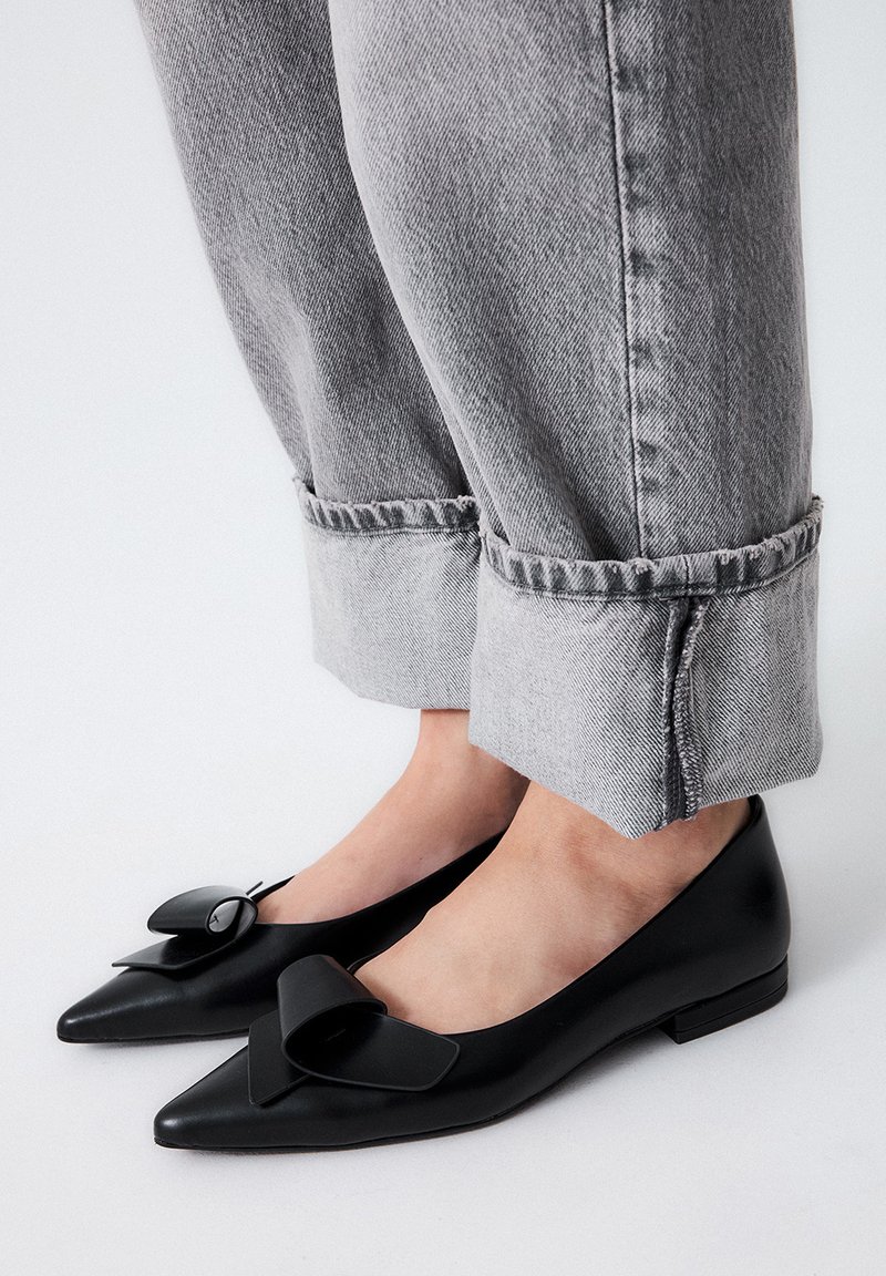 Zapatos planos de cuero negro con puntera afilada, adornados con un gran lazo decorativo en cada zapato. Combinados con unos pantalones vaqueros grises enrollados, que dejan ver los zapatos.