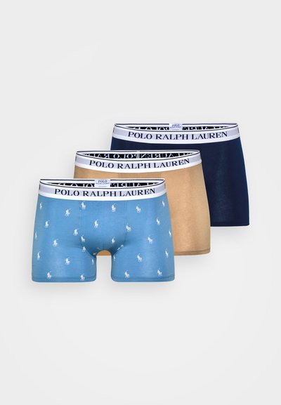 Tri pari moških bokseric: svetlo modre s belimi polo logotipi, bež in mornarsko modre. Vsake so opremljene s črnim pasom z napisom "POLO RALPH LAUREN."