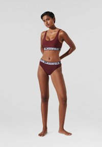 Burgundijas sporta krūšturis un bikinis ar melnbaltu logotipa joslu. Gluda tekstūra, izgriezums ar apaļu kaklu un augsts izgrieziens uz bikini apakšas.