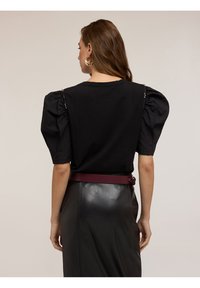 Nainen, jolla on pitkät ruskeat hiukset ja musta puffihihallinen paita, jossa on helmikirjailuja, sekä mustat nahkahousut ja maroonivälinen vyö, kääntynyt poispäin.