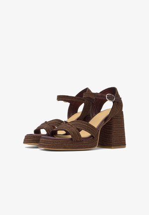 Hnedé pletené platformové sandále s blokovými opätkami a členkovými remienkami so striebornými prackami na bielom pozadí.