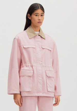 Frau trägt eine hellrosa Jacke mit beigem Kragen und passenden Hosen, steht vor weißem einfarbigem Hintergrund.