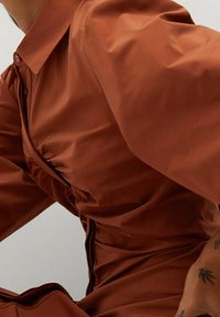Jurkje in roestkleurige stof, gemaakt van glad materiaal, met een kraag, gerimpeld vooraan en lange mouwen met een subtiele textuur.