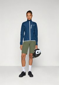 Sötétkék kerékpáros dzseki fehér cipzárral, zöld rövidnadrág, fekete cipők, és fekete-fehér sisak a kézben. Simább anyag, atlétikus szabás.