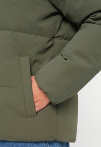 Chaqueta acolchada de color verde oliva con textura de canalé vertical, que incluye un bolsillo lateral y un diseño de manga ajustada. Material suave y aislante.