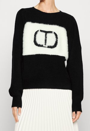 Mujer con suéter negro de felpa con parche rectangular blanco y logo estilizado "CT" en negro, combinado con falda plisada blanca.