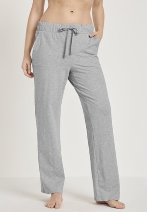 Frau trägt lockere, hellgraue Sweatpants mit elastischem Bund und Kordelzug, steht barfuß da und hat eine Hand in der Tasche.