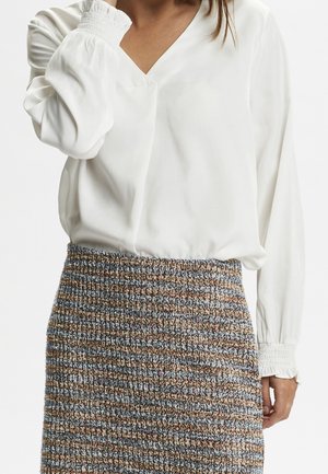 Femme portant un chemisier blanc à manches longues avec poignets froncés et une jupe taille haute texturée aux rayures bleues, marron et beige.