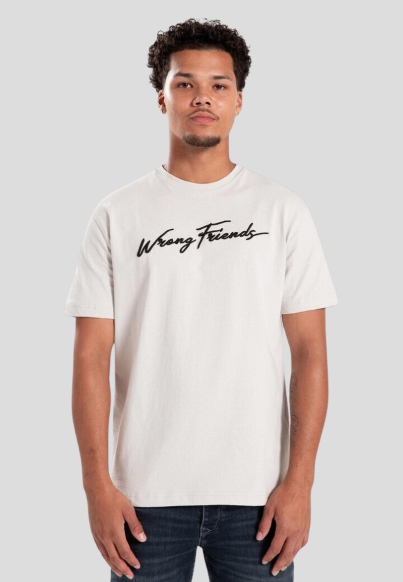 Camiseta de algodón color beige claro con mangas cortas y cuello redondo, que presenta el texto impreso "Wrong Friends" en cursiva negra en el pecho.