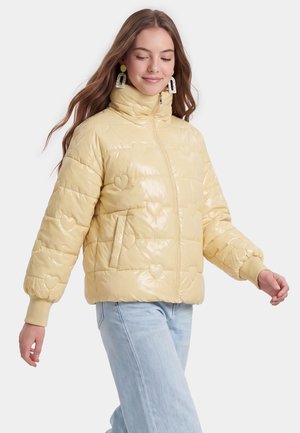 Junge Frau trägt eine hellgelbe gesteppte Daunenjacke mit Herzmustern, hellblaue Jeans und dekorative Ohrringe, geht mit geschlossenen Augen.