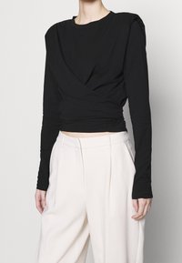 Schwarzes Langarmoberteil mit Wickeldesign und strukturierten Schultern, kombiniert mit hochgeschnittenen, hellbeigen Hosen.