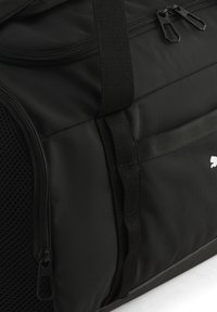 Sort stof sportstaske med netlomme på siden, dobbelte lynlåsvedhæng og et lille hvidt dyrelogo.