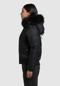 Geacă neagră matlasată, cu glugă detașabilă din blană sintetică, buzunare cu fermoar, manșete tricotate și textură netedă. Vedere laterală care evidențiază detaliile designului.