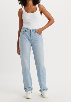 Femme portant un débardeur blanc, un jean taille haute bleu clair et des baskets blanches, debout avec une main dans la poche.