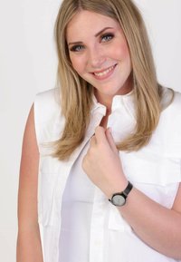 Ronde, zilveren horloge met een zwarte band, gedragen om een linkerpols. De drager heeft een witte mouwloze top aan met twee voorzakken.