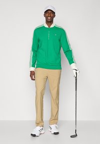 Zelený pulóver na zips po štvrťce s bielym golierom a tromi bielymi pruhmi na rukávoch, skombinovaný s khaki nohavicami a bielymi golfovými topánkami, pričom drží golfovú palicu.