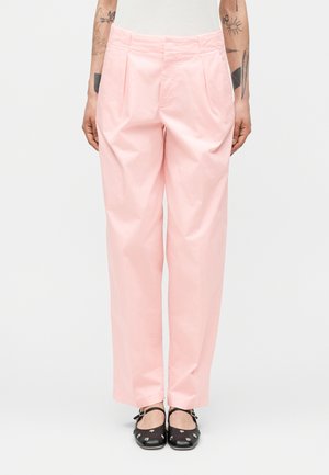 Osoba nosząca jasnoróżowe plisowane spodnie z wysokim stanem oraz czarne buty typu Mary Jane ze srebrnymi oczkami, stojąca na jednolitym tle.