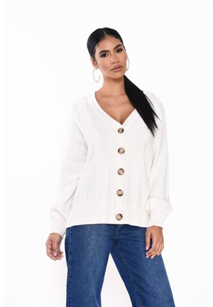 Biała kardigan z plecionki z głębokim dekoltem w kształcie V, z dużymi drewnianymi guzikami i ściągaczami na rękawach. Noszony na niebieskich szerokich dżinsach.