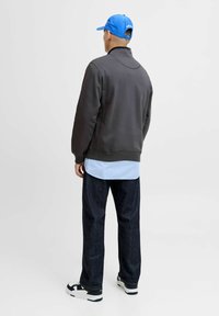 Sudadera gris, gorra azul, camisa a rayas azul claro, jeans oscuros y zapatillas negras con acentos blancos. Conjunto casual visto desde la parte posterior.