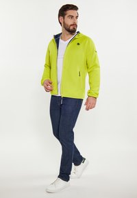Giacca in pile verde lime con zip e cappuccio foderato in blu navy, con dettagli neri e una tasca laterale con zip, abbinata a jeans scuri e sneakers bianche.