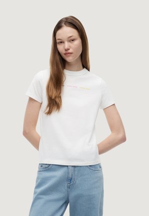 Giovane donna con lunghi capelli castani indossa una maglietta bianca a maniche corte con testo colorato e jeans azzurri, in piedi con le mani dietro la schiena.