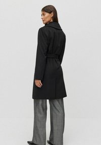 Svart trenchcoat med långa ärmar, bälte i midjan och breda revers; parat med grå vida byxor. Slät textur, knälång design.