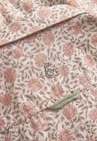 Tessuto a fantasia floreale in un delicato rosa, con intricate fiori verdi e arancioni. Include un anello in tessuto verde e un'emblema ricamato.
