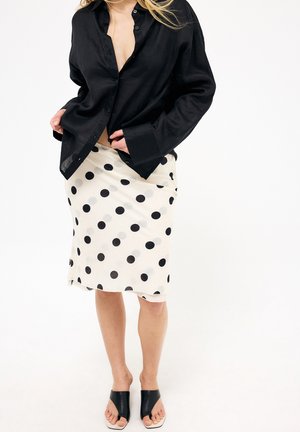 Osoba ubrana w czarną koszulę z guzikami, białą spódnicę do kolan z dużymi czarnymi kropkami oraz czarne sandały na obcasie z odkrytymi palcami.