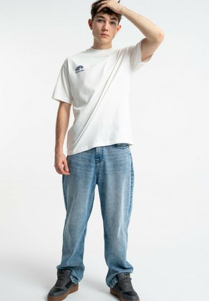 Młody mężczyzna w białej koszulce z nadrukiem, luźnych niebieskich dżinsach i czarnych trampkach, stojący z jedną ręką na głowie na tle jednolitej białej ściany.