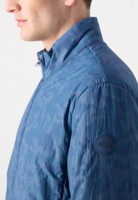 Casaco de camuflagem azul marinho com um acabamento texturizado, apresentando um colarinho de pé e um emblema circular no braço esquerdo.