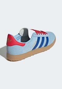 Baskets en suede bleu clair avec des lacets et un talon rouges, comportant trois rayures bleu marine et une semelle en caoutchouc beige. Logo doré sur le talon.