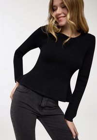 Top nero in maglia a coste con maniche lunghe e orlo svasato, abbinato a jeans grigio scuro slim-fit su sfondo chiaro.