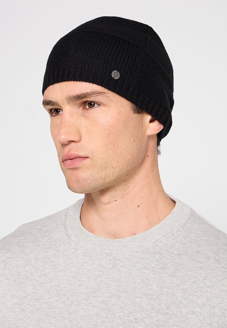 Młody mężczyzna w czapce typu beanie w kolorze czarnym i jasnoszarym swetrze z okrągłym dekoltem, spoglądający lekko w lewo na jednolitym tle.