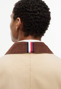 Άτομο με κοντά σγουρά μαλλιά που φοράει μπεζ μπουφάν με καφέ γιακά από κοτλέ, με μια ρίγα κόκκινου, λευκού και μπλε στη μέση.