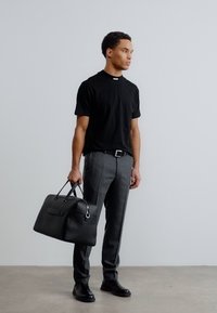 Άνδρας που φοράει μαύρο μπλουζάκι και γκρι παντελόνι, κρατώντας μια μαύρη δερμάτινη τσάντα duffel, στέκεται μπροστά σε απλή ανοιχτό γκρι επιφάνεια τοίχου.