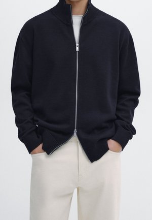 Osoba w czarnym swetrze rozpinanym na zamek, na białej koszuli, z rękami w kieszeniach kremowych spodni, na jednolitym tle.