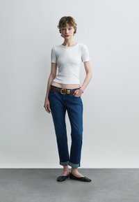 Young person standing with hand in pocket, wearing a white fitted short-sleeve top, blue rolled-up jeans, brown belt, and black flats.