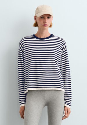 Jeune personne portant une casquette beige, un t-shirt à manches longues rayé bleu marine et blanc, et un pantalon gris clair sur un fond clair uni.