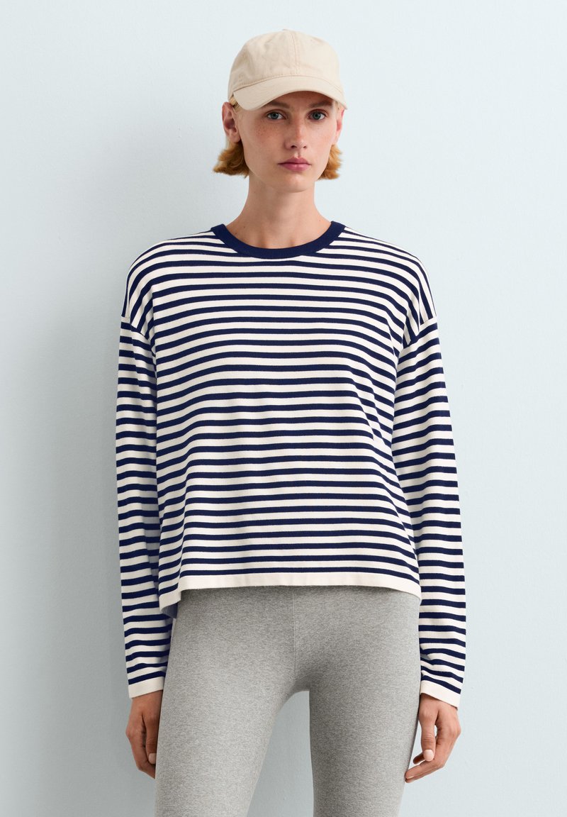 Jeune personne portant une casquette beige, un t-shirt à manches longues rayé bleu marine et blanc, et un pantalon gris clair sur un fond clair uni.
