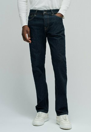 Hombre con vaqueros de pierna recta azul oscuro, camisa blanca de manga larga y zapatillas blancas, de pie con una mano en el bolsillo.