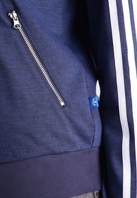 Námořnická zipová mikina s texturovaným materiálem, bílými postranními pruhy a modrou logovou nášivkou umístěnou v dolní levé části blízko zipu.