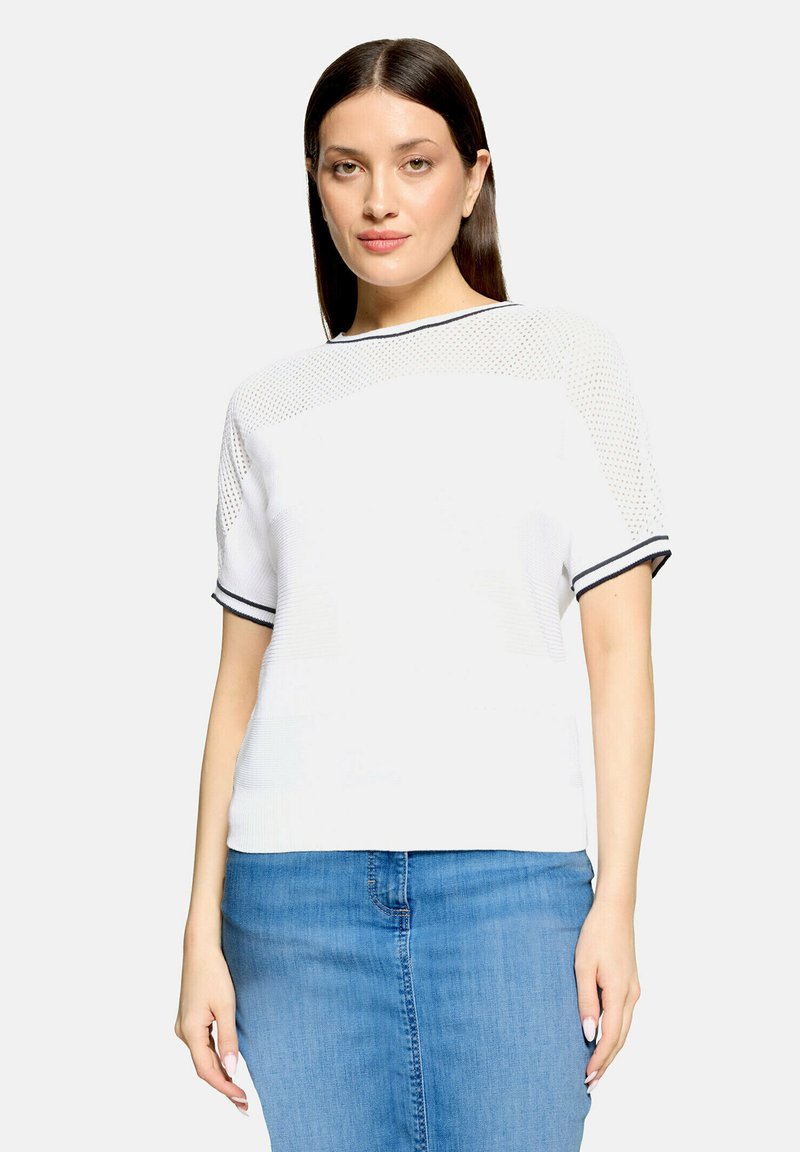 Femme aux longs cheveux bruns portant un haut en tricot blanc à manches courtes avec un bord noir et une jupe en jean bleue, debout devant un fond uni.