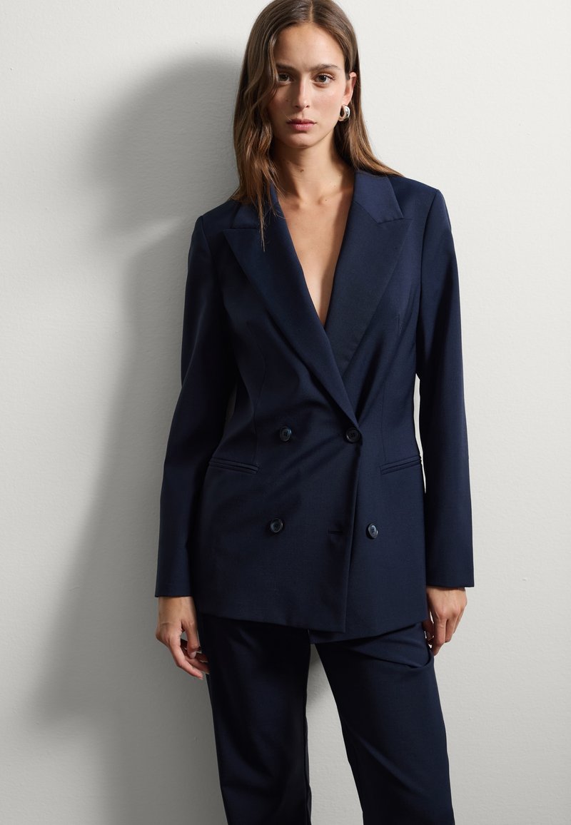 Donna che indossa un blazer doppiopetto blu navy e pantaloni coordinati, in piedi contro un muro bianco semplice, con lunghi capelli castani e orecchini a cerchio.