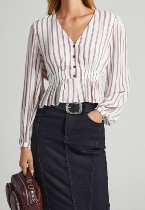 Femme portant un chemisier blanc à rayures rouges verticales et boutons, rentré dans une jupe noire avec une large ceinture, tenant un sac à main bordeaux.