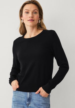 Woman wearing a black long-sleeve sweater and blue jeans, standing with hands in pockets against a plain light background.