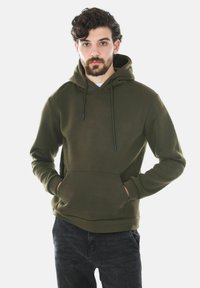 Felpa verde oliva con tasca frontale, cordini regolabili e polsini a coste. Il tessuto appare liscio con una vestibilità casual. Indossata sopra jeans scuri.