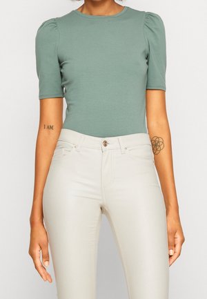 Mujer que lleva una camiseta ajustada de manga corta verde salvia con hombros abullonados y pantalones crema de talle alto con cierre de botones.
