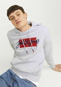 Grijze hoodie van zacht materiaal met een voorzak, voorzien van een rode en navy logo print. Een witte laagshirt en blauwe jeans zijn zichtbaar.