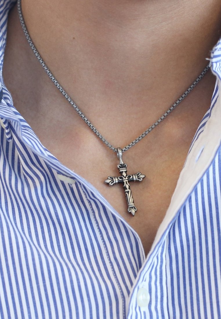 Silberfarbener Kreuzanhänger an einer Kette, mit filigranen Designs und floralen Akzenten, getragen über einem blau-weiß gestreiften Hemd.
