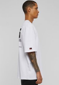 Balts kokvilnas T-krekls ar lielu ieguldījumu, ar drukātu tekstu aizmugurē, īsām piedurknēm un mazu logotipa uzlīmi uz piedurknes.